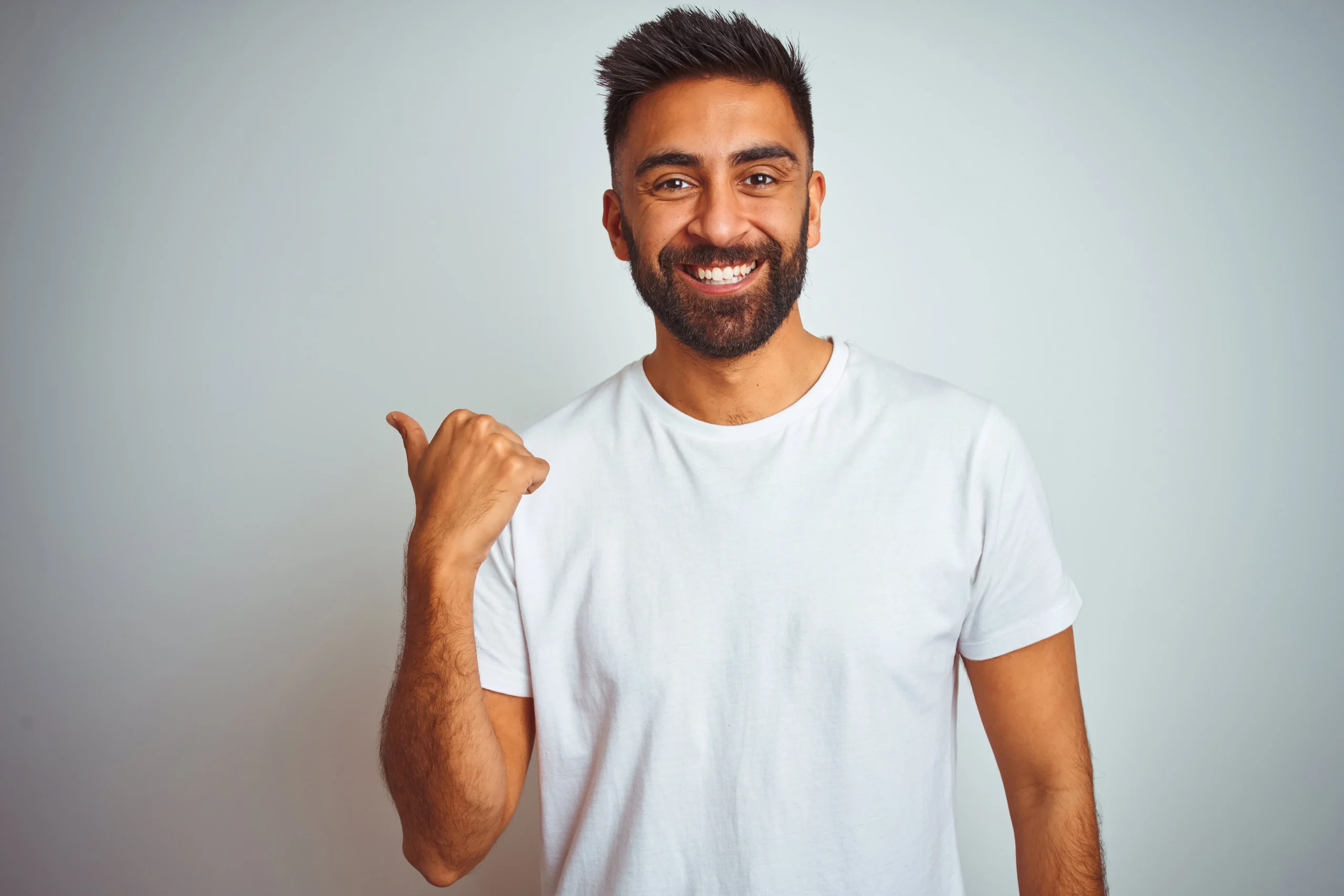 Smiling man in a white t-shirt pointing his thumb over his shoulder against a plain background.