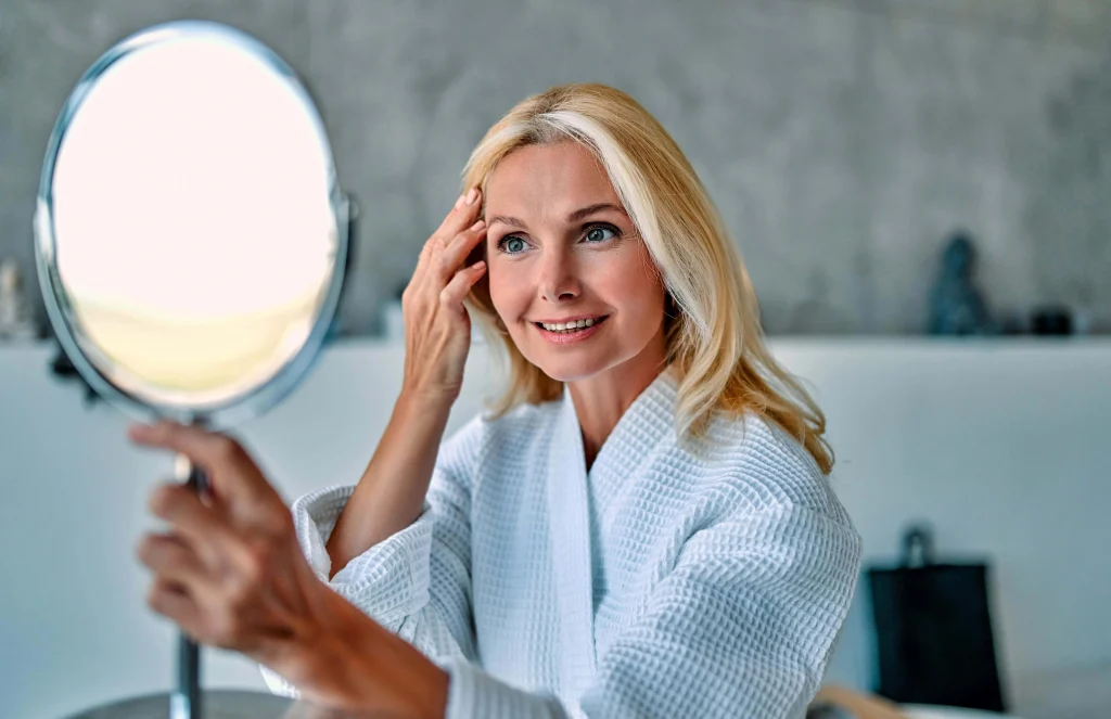 Smiling woman in a white robe looking in a handheld mirror and touching her face.