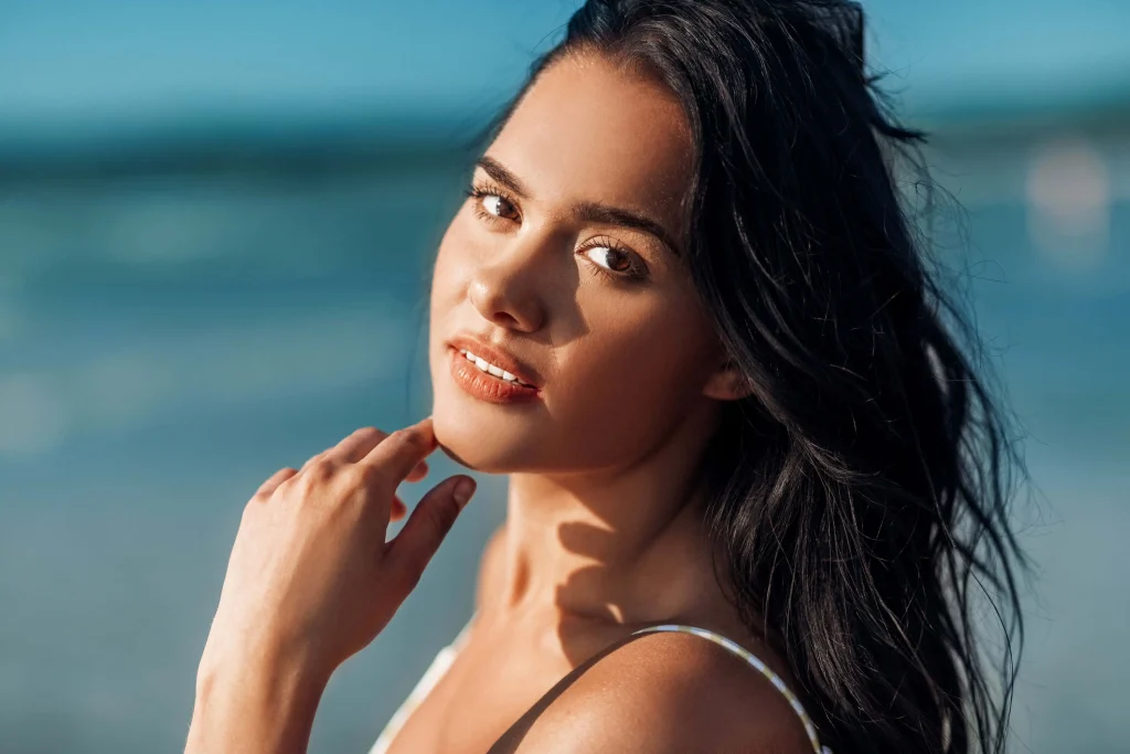 Woman with long dark hair posing on the beach, looking at the camera with a soft smile.