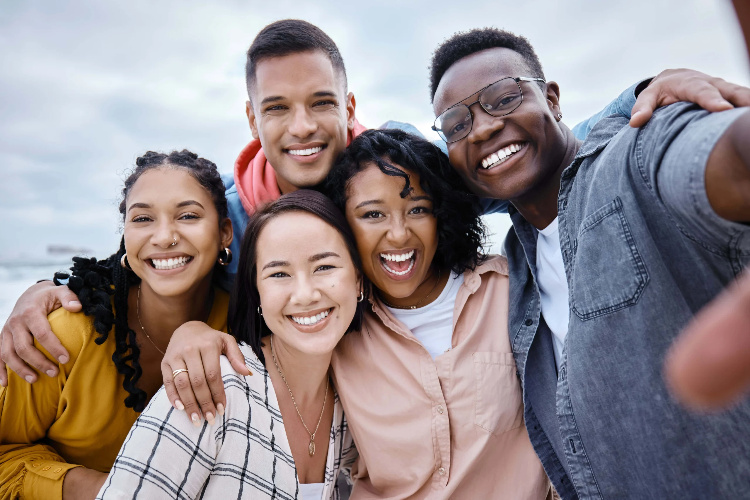 Five smiling friends pose closely together at the beach, showcasing joy and camaraderie.