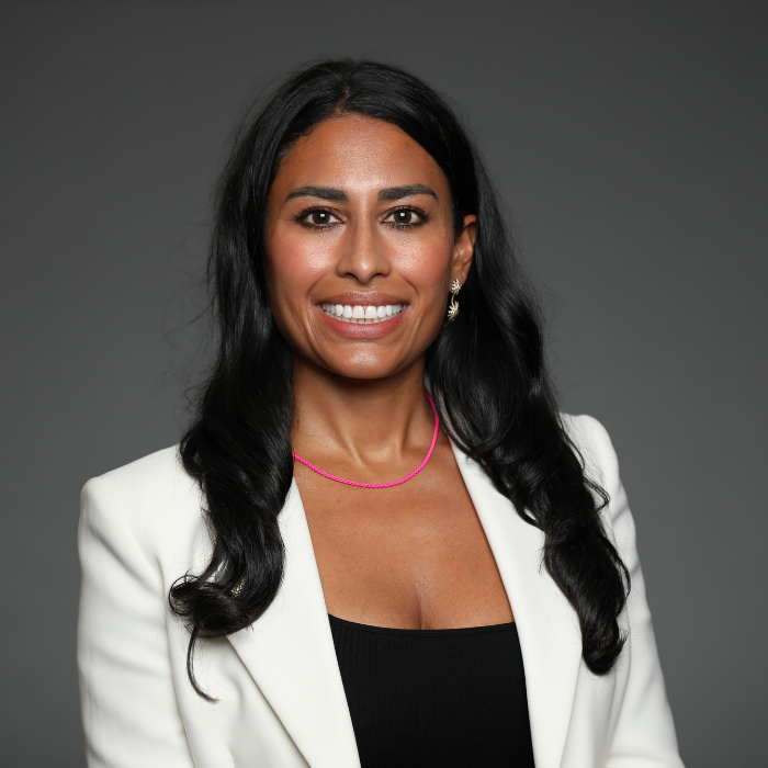 Woman with long black hair, wearing a white blazer and pink necklace, smiling against a gray background.